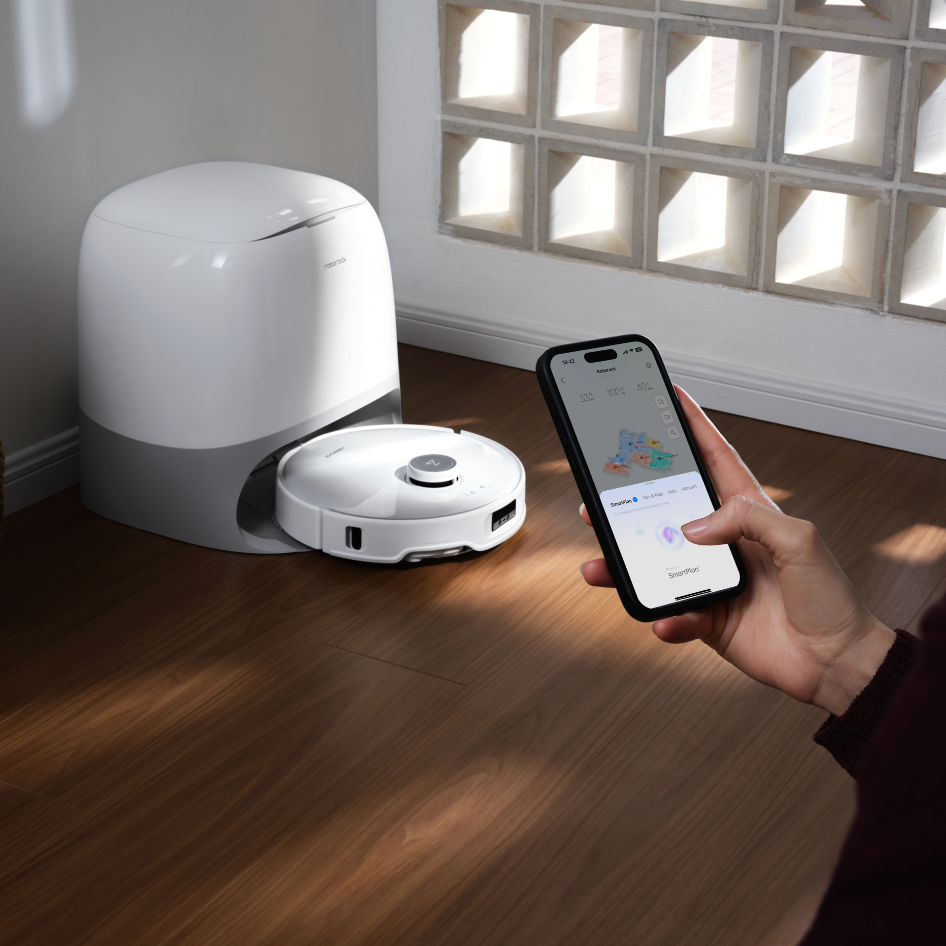 White Roborock robot vacuum docked in a white base station on a wooden floor. In the foreground, a hand holds a smartphone displaying a cleaning map on the Roborock app