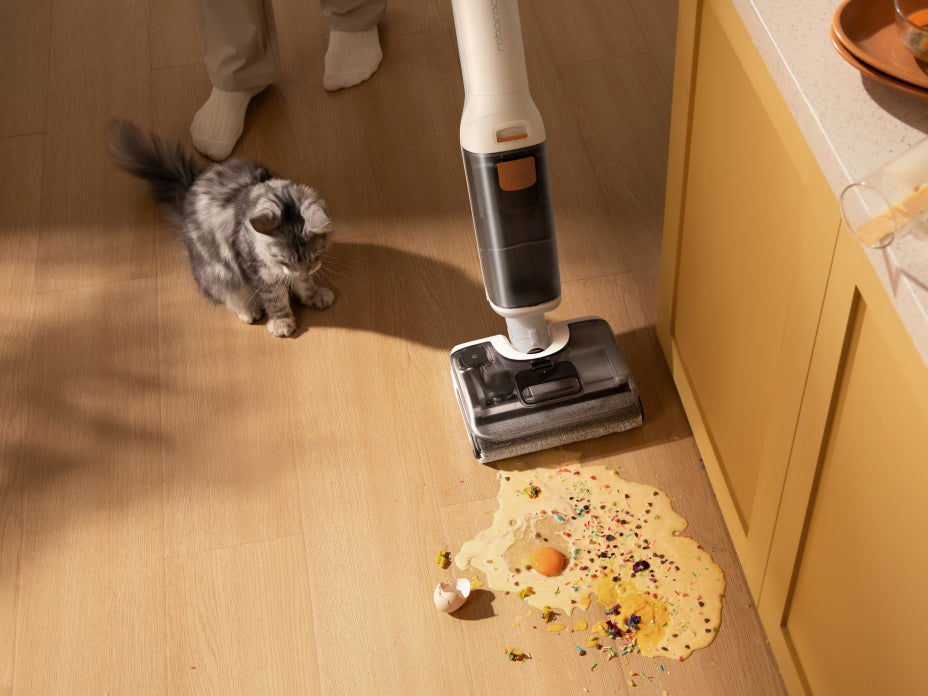 A person using a white Roborock F25 GT wet/dry vacuum to clean up a cracked egg and spill on a wooden kitchen floor, with a small kitten watching nearby