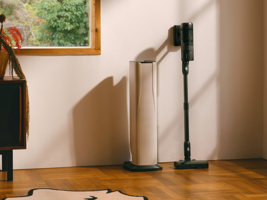 Black Roborock H60 Hub Ultra cordless stick vacuum leaning against a white wall next to its docking station on a wooden floor