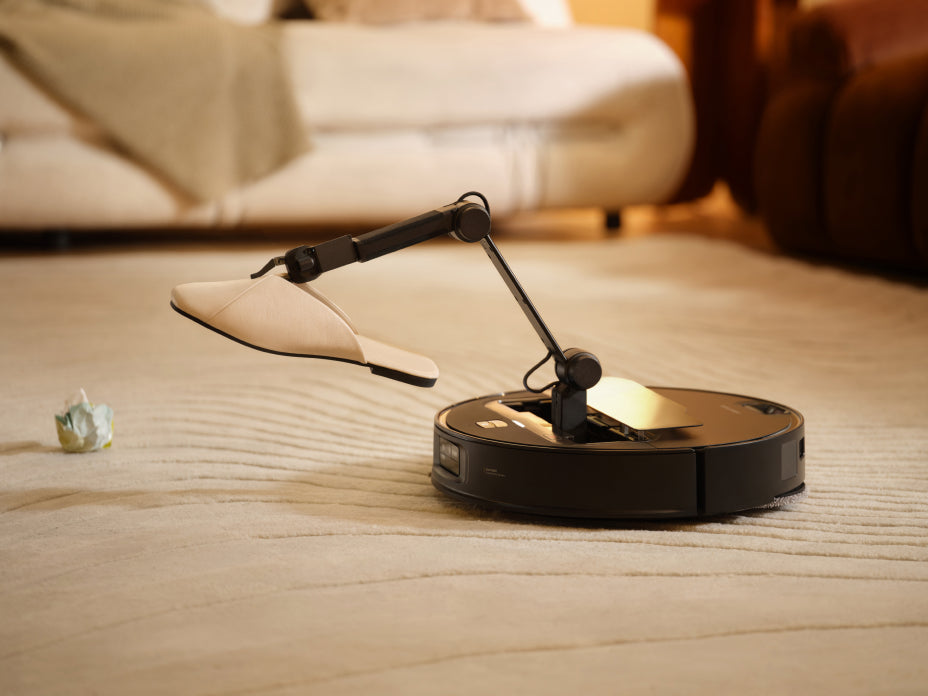 Black Roborock Saros Z70 robot vacuum on a rug, using an extended mechanical arm to lift a slipper